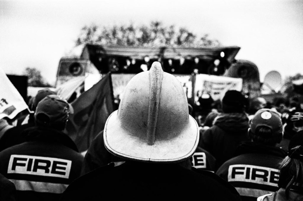 “FAIR PAY IS 30K” Fire Brigades Union (FBU) demonstration, Hyde Park, London, 2002. In December 2002, 20,000 firefighters and trade unionists from around the UK marched through the centre of London demanding fairer pay for British firefighters. As night fell over the city and the demonstration drew to a close, the crowd heard Fire Brigades Union General Secretary Andy Gilchrist “douse hopes of a swift end to the dispute with a combative speech warning that the union would pull out of mediation talks at the first sign of Government intervention” (The Guardian). Image copyright © Thaddeus Pope 2002.