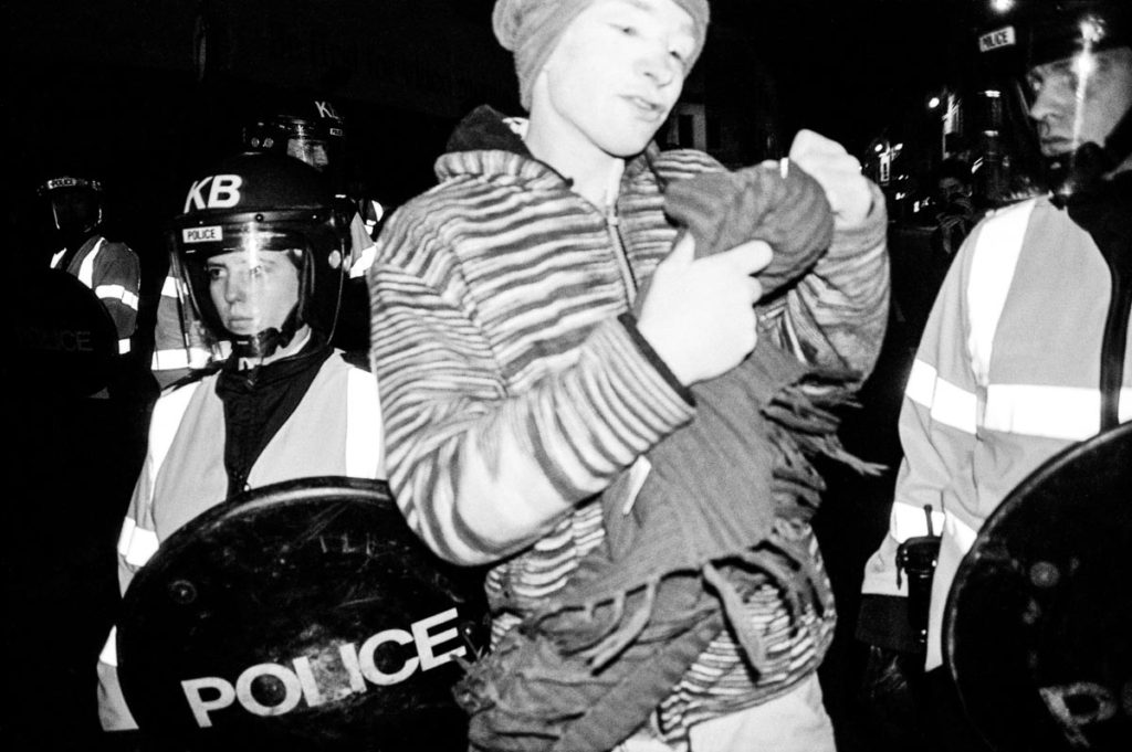 An anti-war protestor wearing white face paint dances through a line of riot police on Brighton Seafront, England (2003). Image © Thaddeus Pope