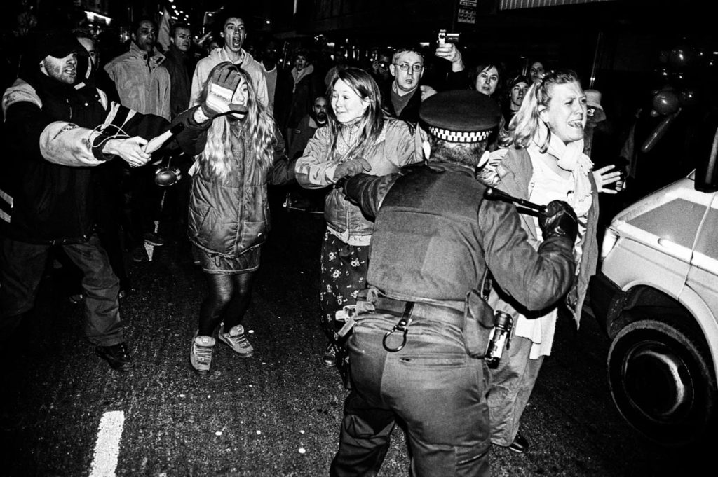 Armed with a baton, a British policeman uses force to control crowds of anti-war protestors in Brighton, England (2003). Image © Thaddeus Pope. (1/2)