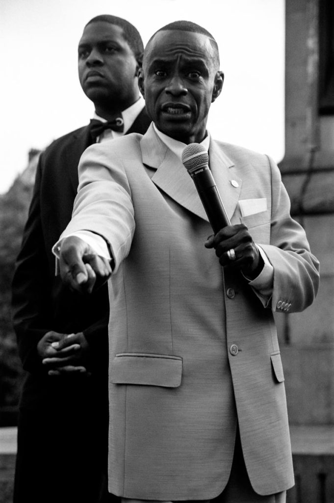 Standing on a platform in Trafalgar Square against the backdrop of Nelson's Column, minsters from the Nation of Islam address a large crowd of their followers during the Million Man March, London, England, 2004. Image copyright © Thaddeus Pope 2004.